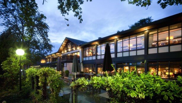 Un hôtel moderne doté de grandes fenêtres, d’un espace extérieur aménagé et d’une végétation luxuriante.
