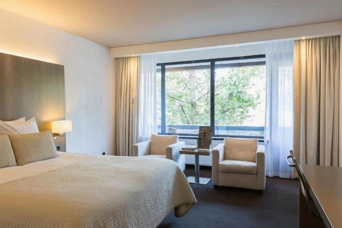 A hotel room with a large bed, a desk, two chairs, and a window with curtains.