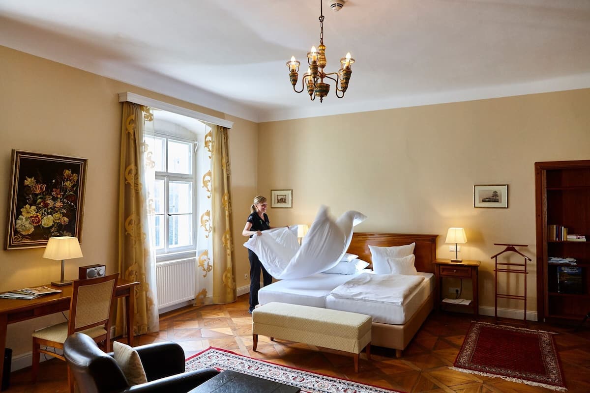 A hotel room with a bed, a desk, a chair, a window with curtains, a chandelier, and a bookshelf.