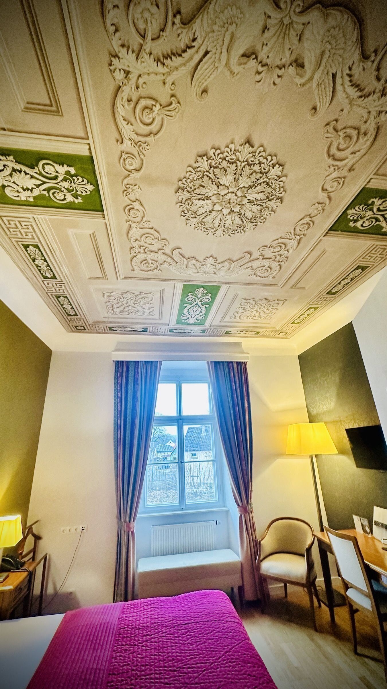 Ein Zimmer mit Bett, Schreibtisch, Stuhl, einem Fenster mit Vorhängen und aufwendigen Details an der Decke.