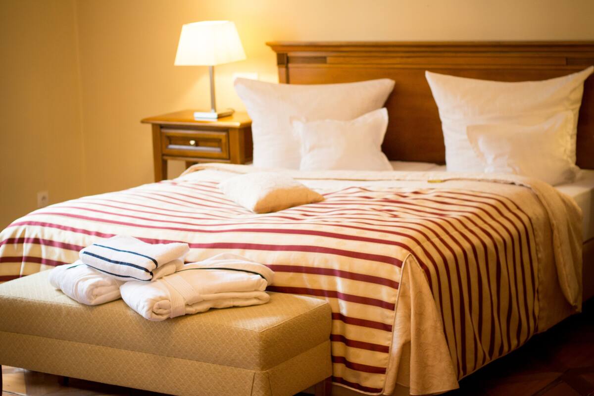 A hotel room with a bed, a nightstand with a lamp, and a bench with towels.