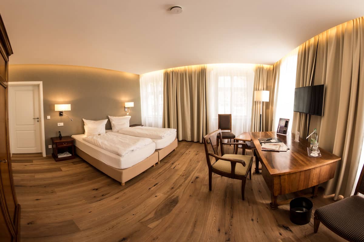 A hotel room with a wooden floor, a bed, a desk, a chair, and a TV.