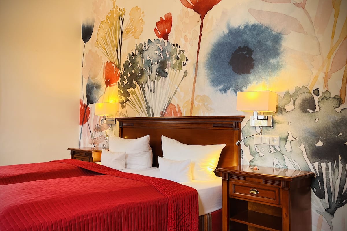 A hotel room with a large bed, a nightstand, a lamp, and a floral mural on the wall.