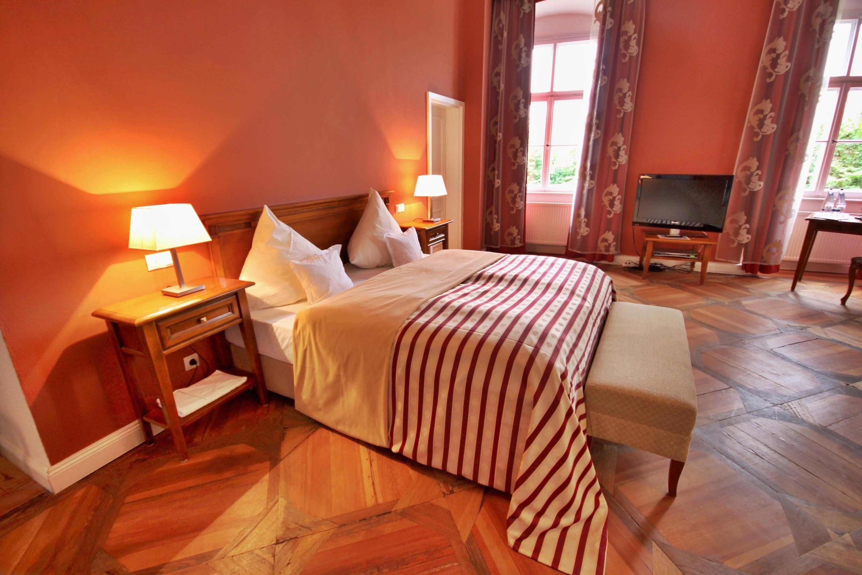Une chambre d’hôtel avec un grand lit, deux tables de chevet avec des lampes, un téléviseur posé sur un meuble et une vue sur l’extérieur par une fenêtre avec des rideaux.