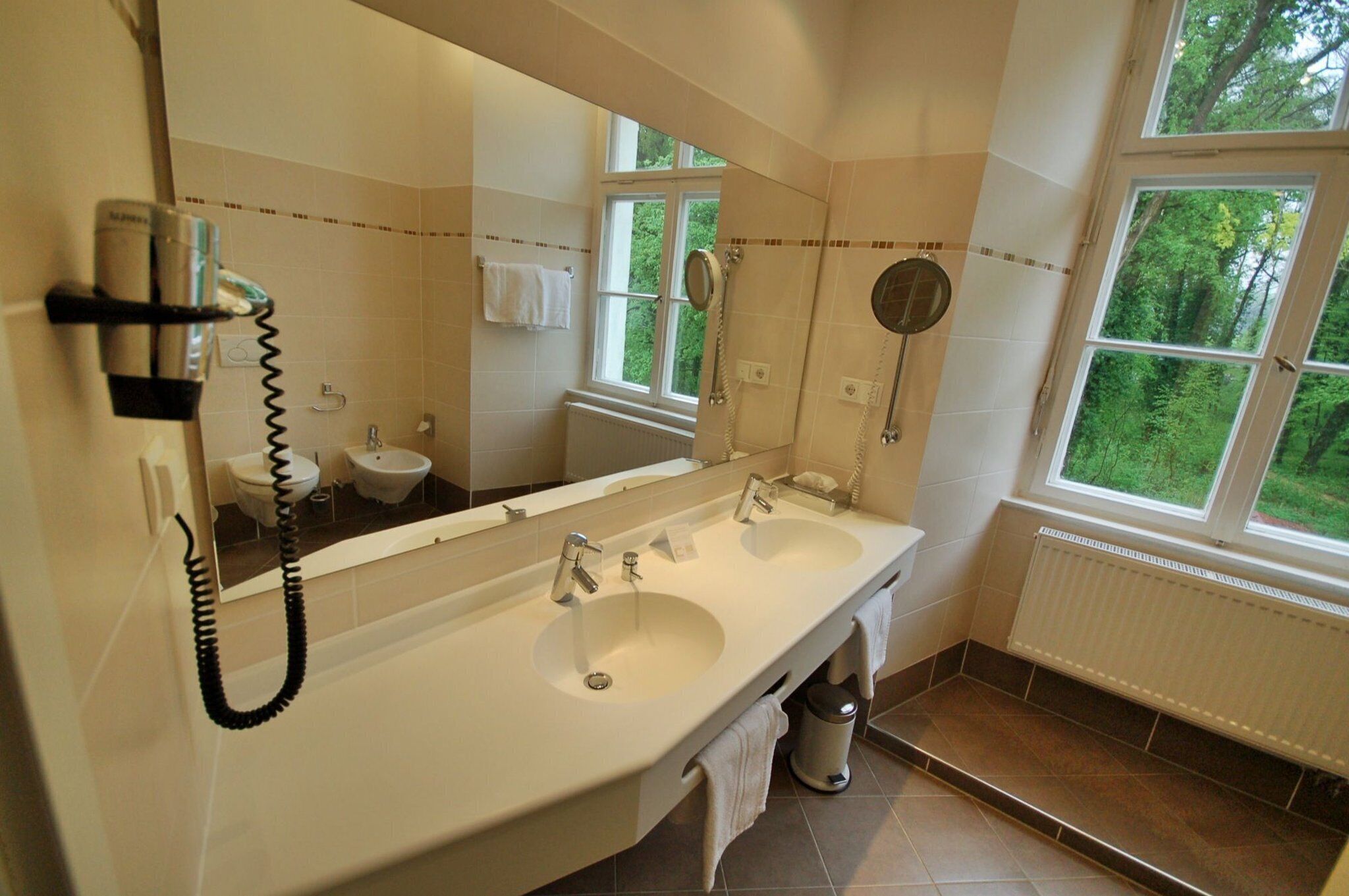 A bathroom with two sinks, a large mirror, a hairdryer, and a window with a view of greenery.