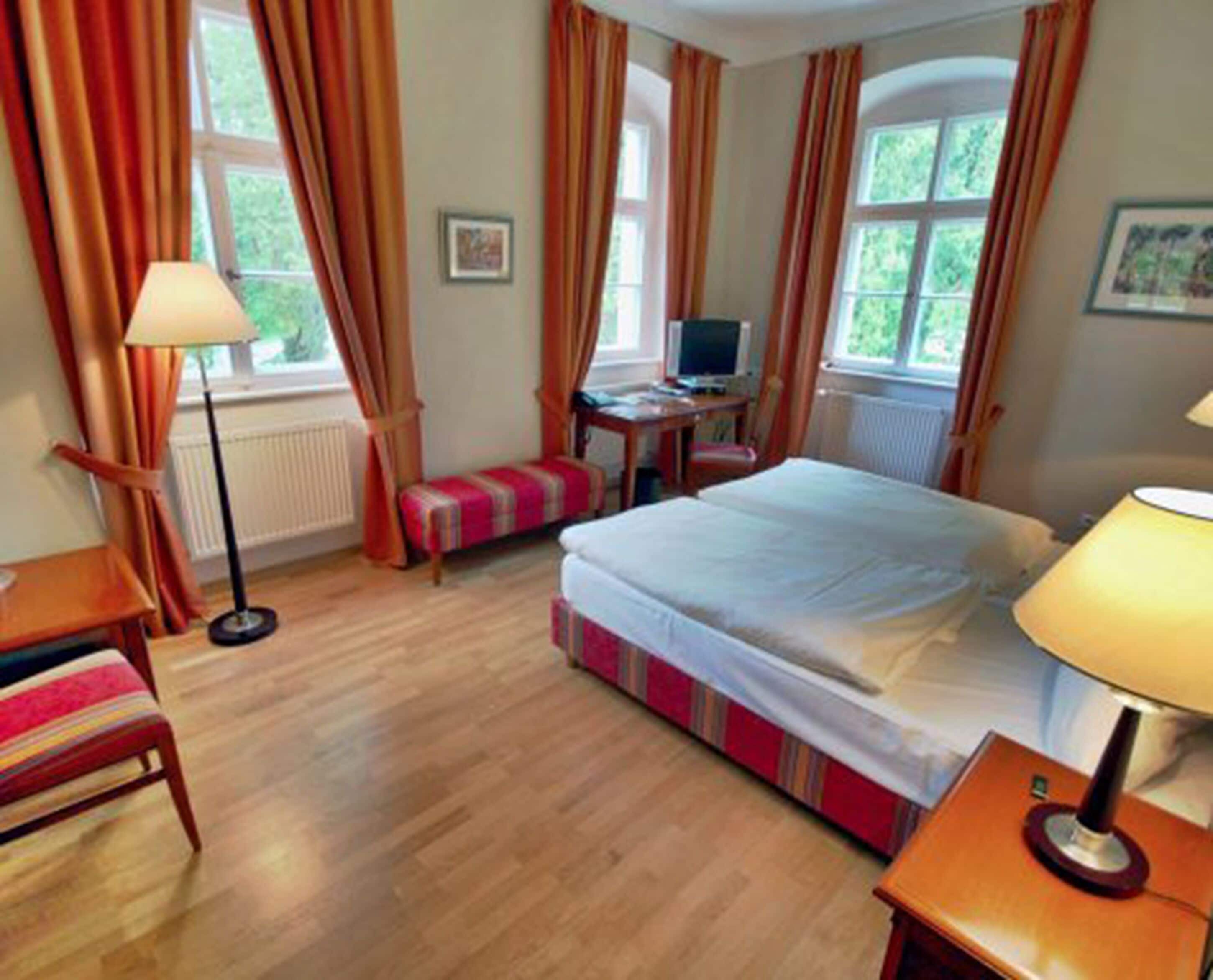 Habitación de hotel con cama, escritorio, silla, lámpara y ventana con cortinas.