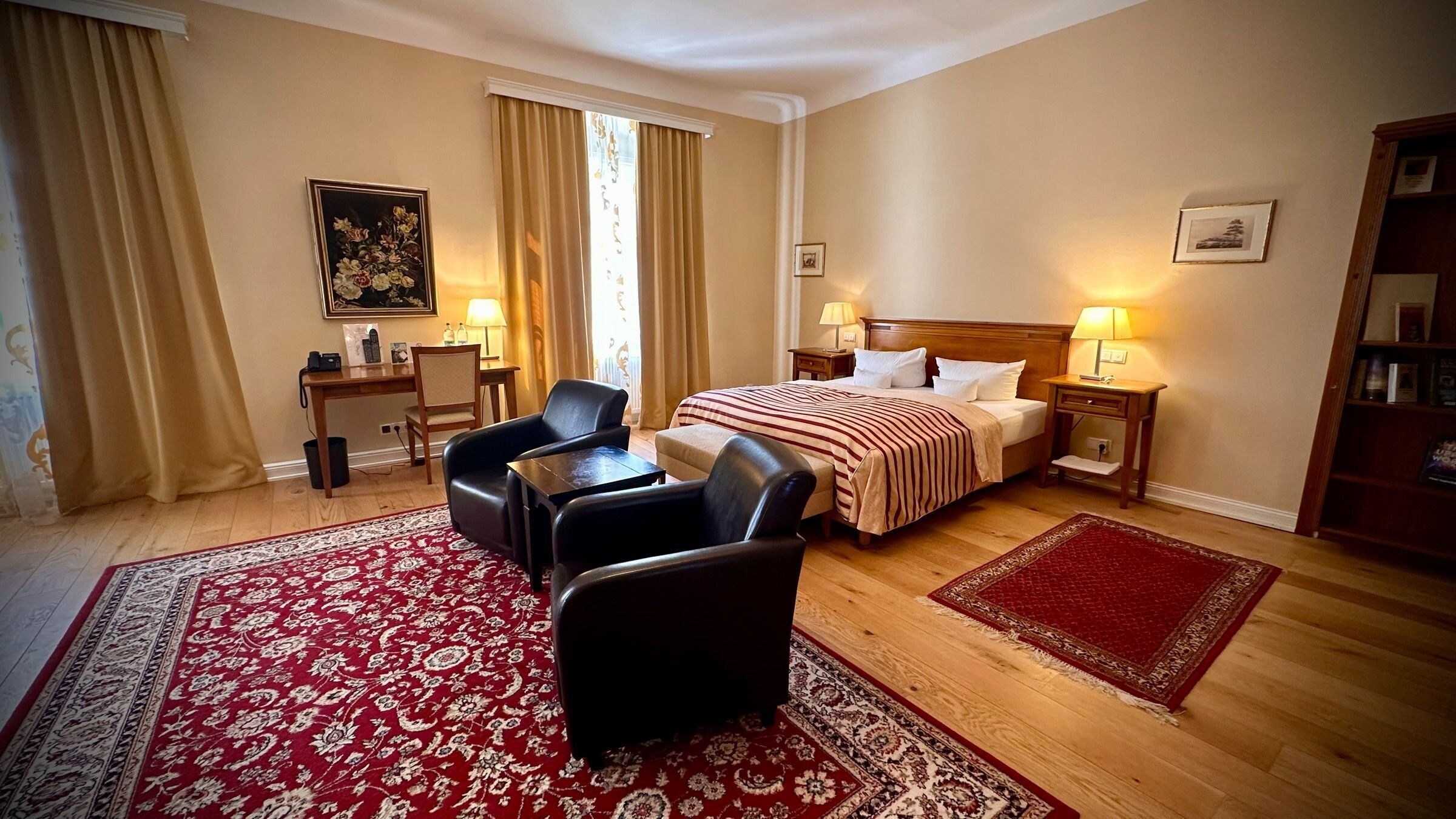 A hotel room with a bed, two bedside tables with lamps, a desk with a chair, a small bookshelf, and a large red and white patterned rug.