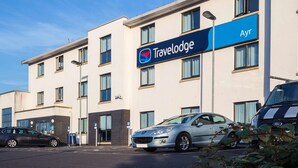 A Travelodge hotel with a modern design, featuring a prominent blue sign and a parking area with several cars. - Travelodge Ayr (Ayr)