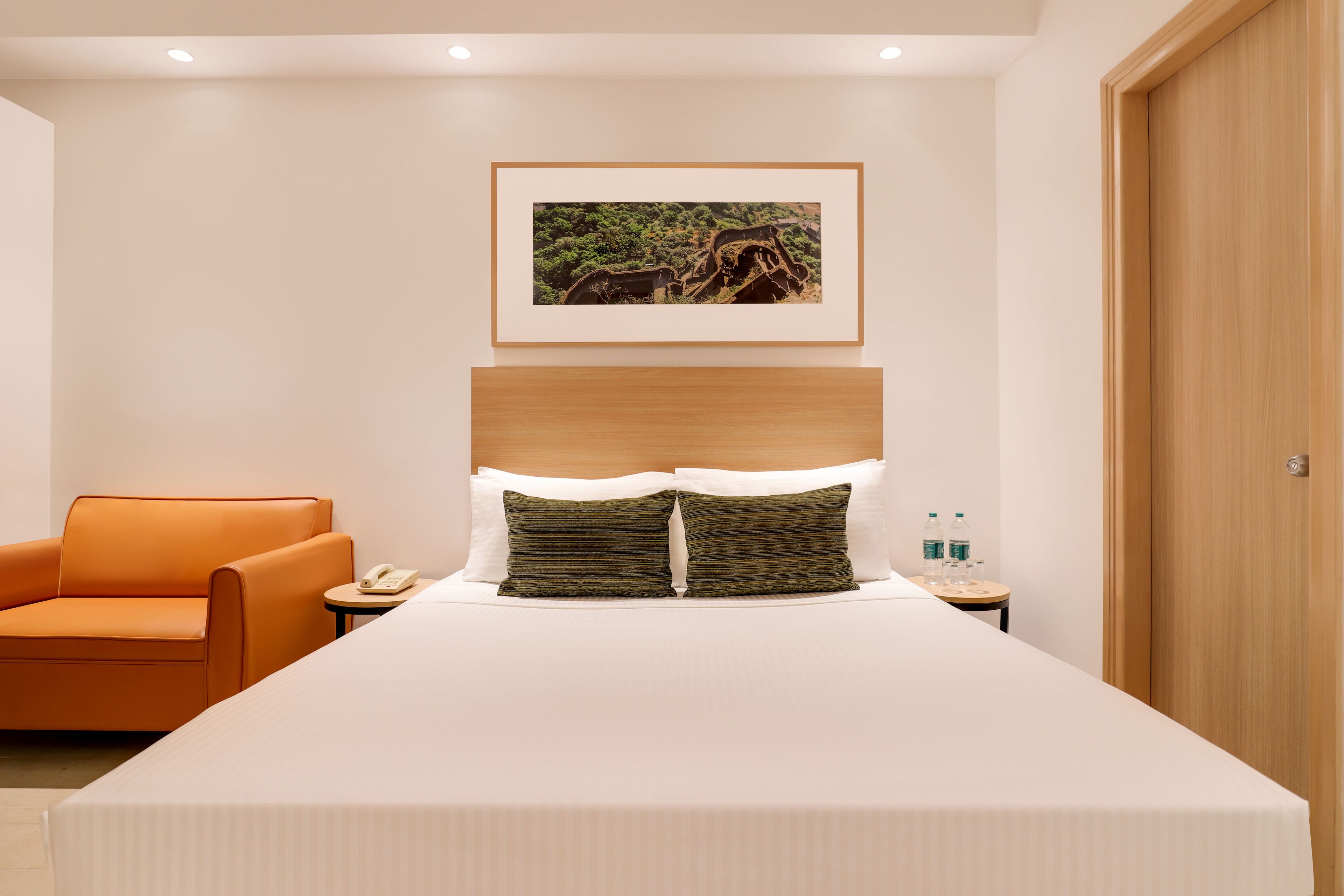 Habitación de hotel con una cama grande, cabecera de madera, un cuadro enmarcado en la pared, una silla naranja y una mesita de noche con un teléfono y botellas de agua.