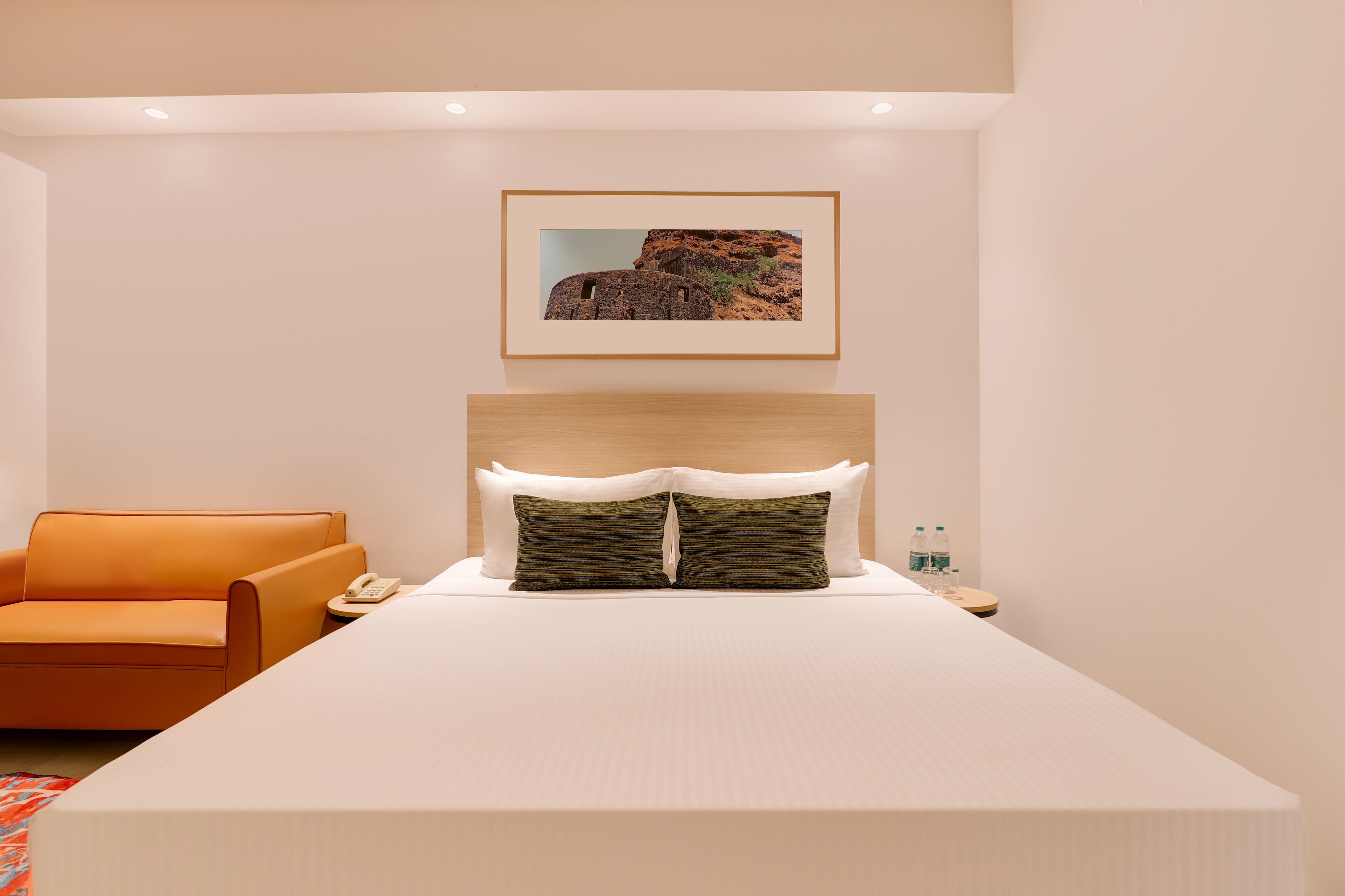 A modern hotel room with a large bed, a wooden headboard, a framed picture on the wall, a small table with a telephone and water bottles, and an orange armchair.