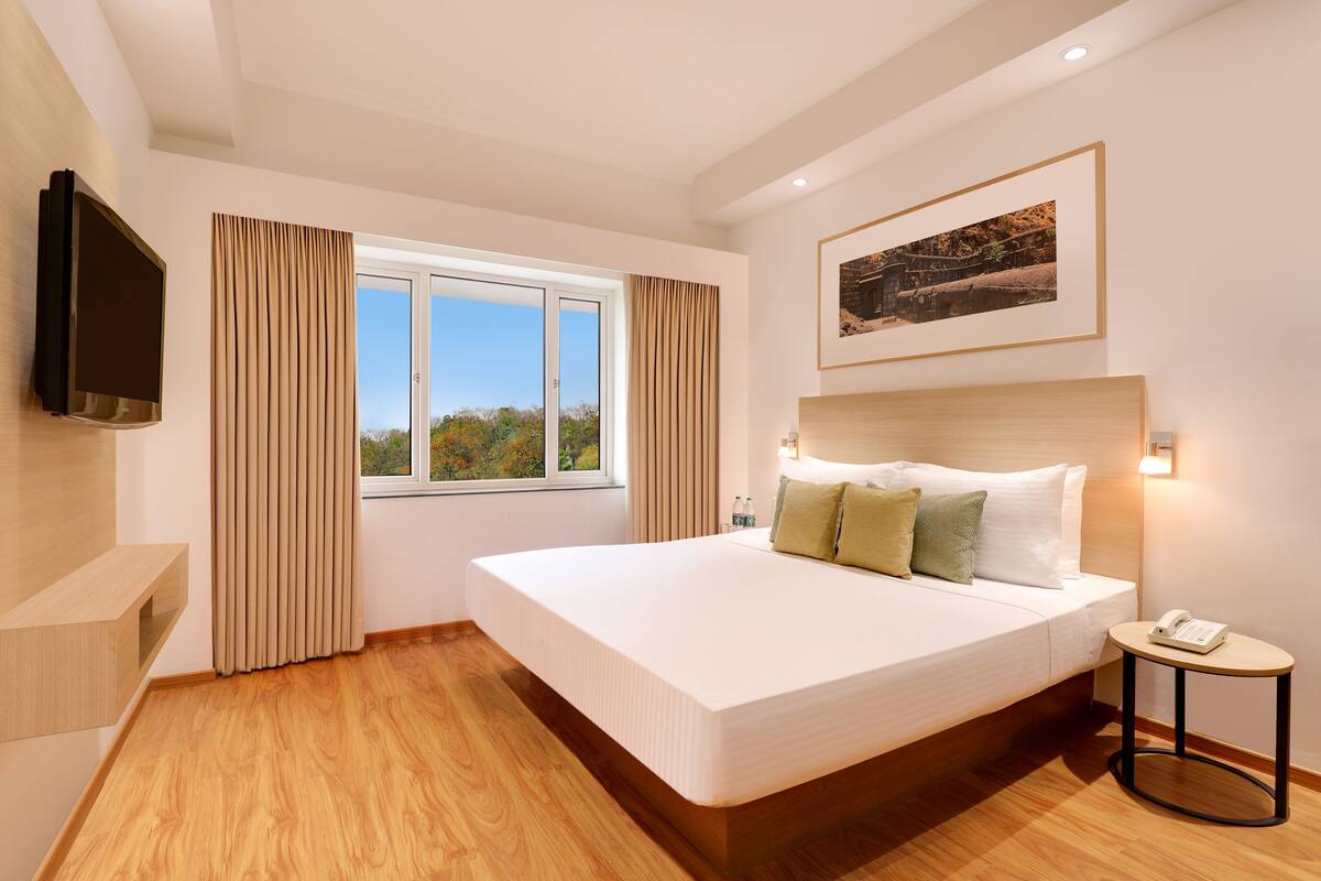 A modern hotel room with a large bed, a flat-screen TV, a bedside table with a phone, and a window with curtains.