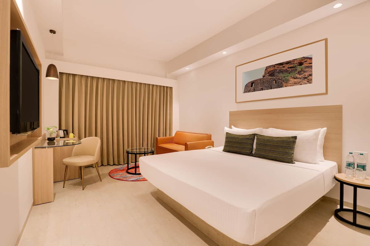 A hotel room with a large bed, a desk with a chair, a small table with a chair, and a TV on the left wall.