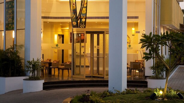 L’entrée d’un hôtel au design moderne, caractérisée par une façade en verre, une colonne décorative centrale et de nombreuses suspensions lumineuses.