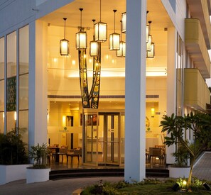 A hotel entrance with a modern design, featuring a glass facade, a central decorative column, and multiple hanging lights.