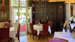 A dining area with wooden tables and chairs, a large window with a view of greenery, and a dark wooden cabinet with glass doors. - Hotel - Restaurant Kronprinz (Falkensee)