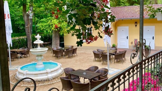 An outdoor patio area with a fountain, wicker furniture, and a yellow building with white doors. - Hotel - Restaurant Kronprinz (Falkensee)