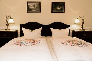 Una cama doble con cubrecamas florales, dos mesitas de noche con lámparas, y dos cuadros en la pared.