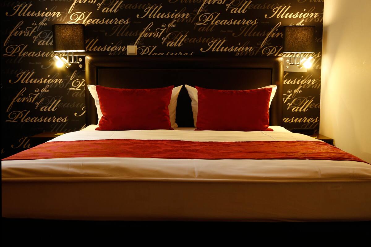 A hotel room with a large bed, red and white pillows, and a wall with repeated text.