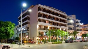 Un hotel de varios pisos con balcones, un estacionamiento y una calle con coches y peatones.
