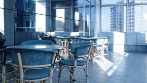 A modern indoor dining area with round tables and wicker chairs, large windows offering views of buildings, and a tiled floor. - Park Hotel (Dnipro)