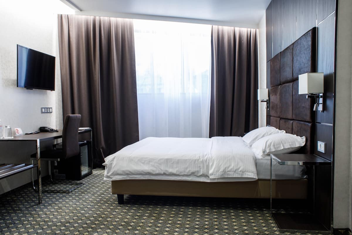 Ein Hotelzimmer mit einem Bett, einem Schreibtisch mit Stuhl, einem Fernseher und einem großen Fenster mit Vorhängen.
