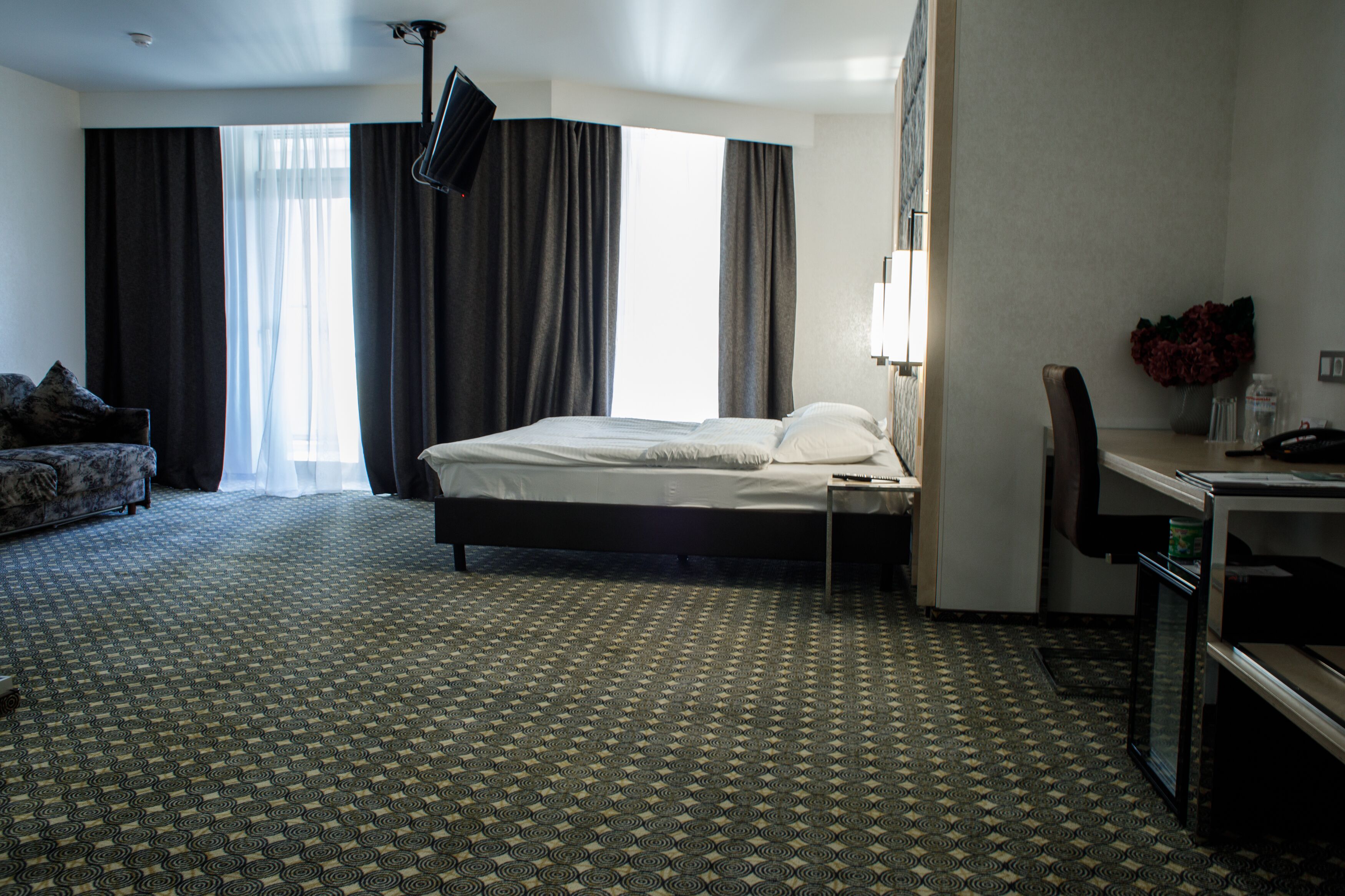 Habitació d'hotel amb un llit, escriptori, cadira, taula petita, mirall, finestra amb cortines i una catifa estampada.