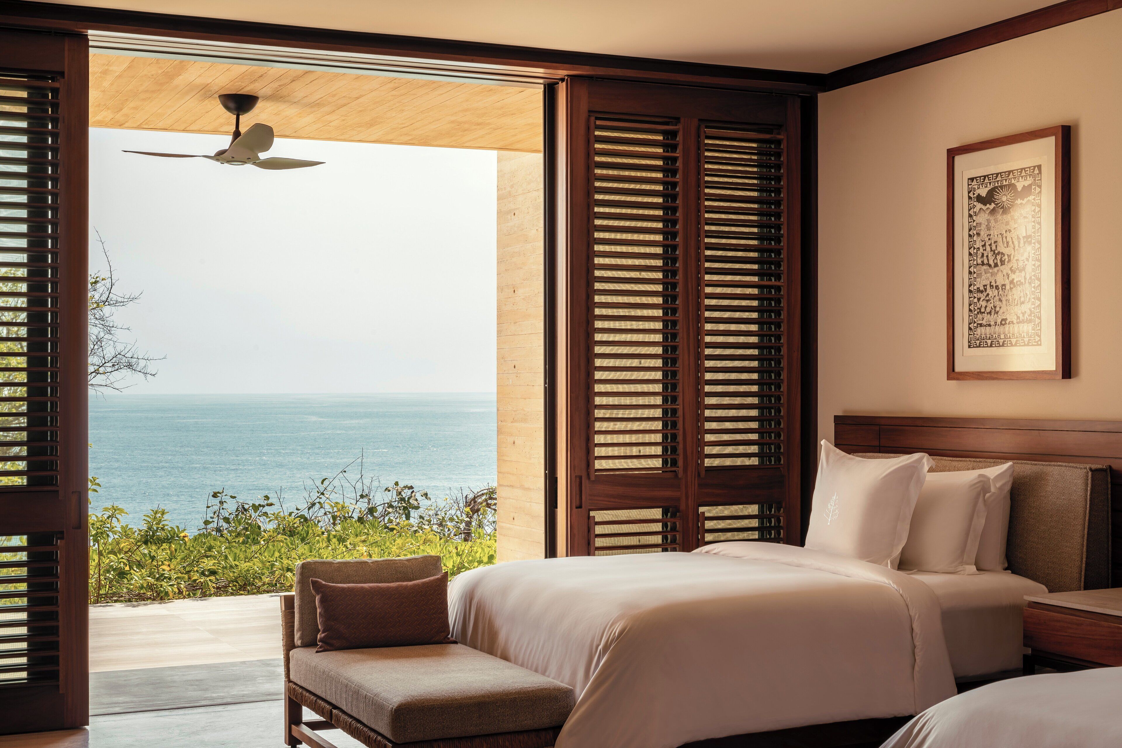 A bedroom with a large bed, a smaller bed, a nightstand, and a chair. There is a window with wooden shutters and a view of the ocean.
