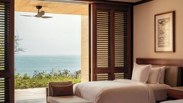 A bedroom with a large bed, a smaller bed, a nightstand, and a chair. There is a window with wooden shutters and a view of the ocean.