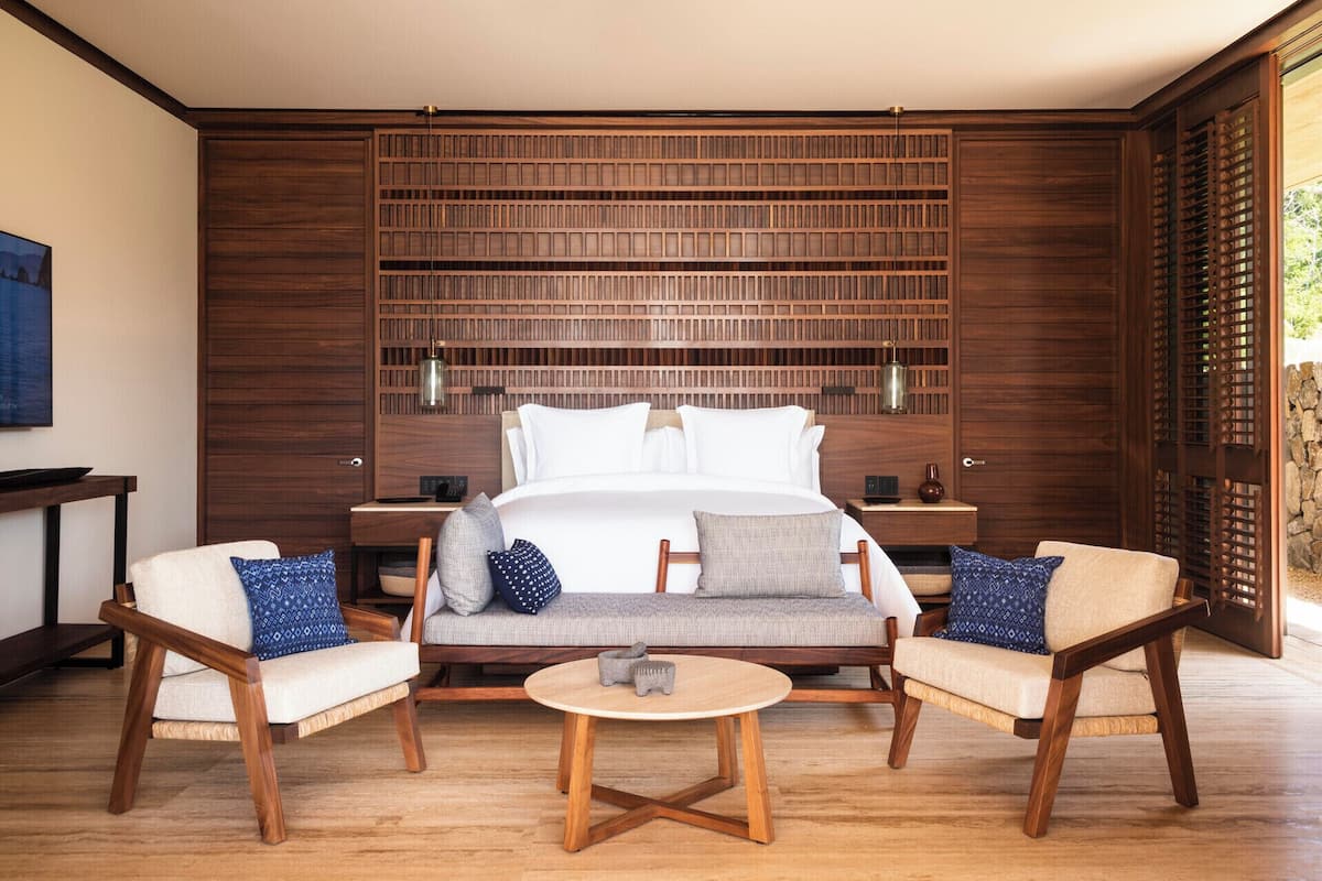 Une chambre moderne avec un grand lit, une tête de lit en bois, deux tables de chevet, un canapé, deux fauteuils, une table basse et une vue sur la verdure par une fenêtre.