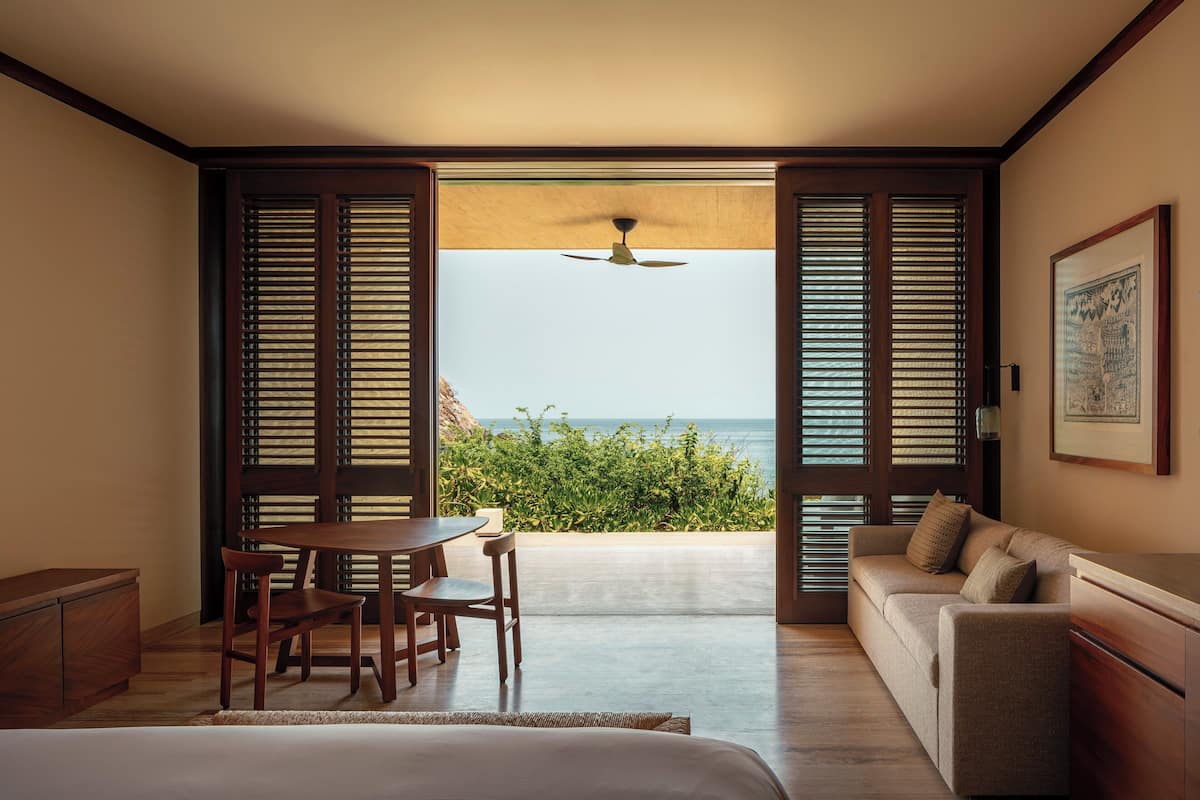 Une chambre avec un lit, un bureau en bois, une chaise, un canapé et une vue sur l’océan à travers des volets ouverts.