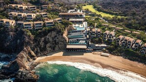 Vista aérea de un complejo turístico en la costa con varios edificios, una playa y una zona de piscina.