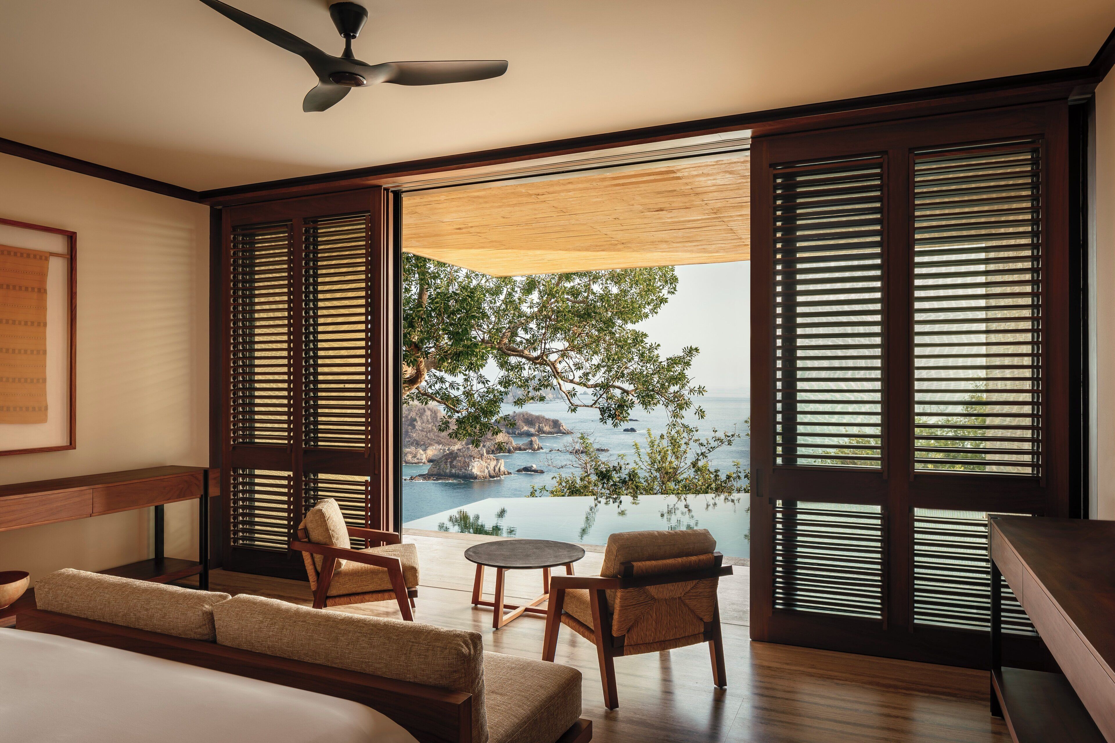 A modern hotel room with a large sliding glass door leading to a balcony with a view of the sea and rocky landscape.
