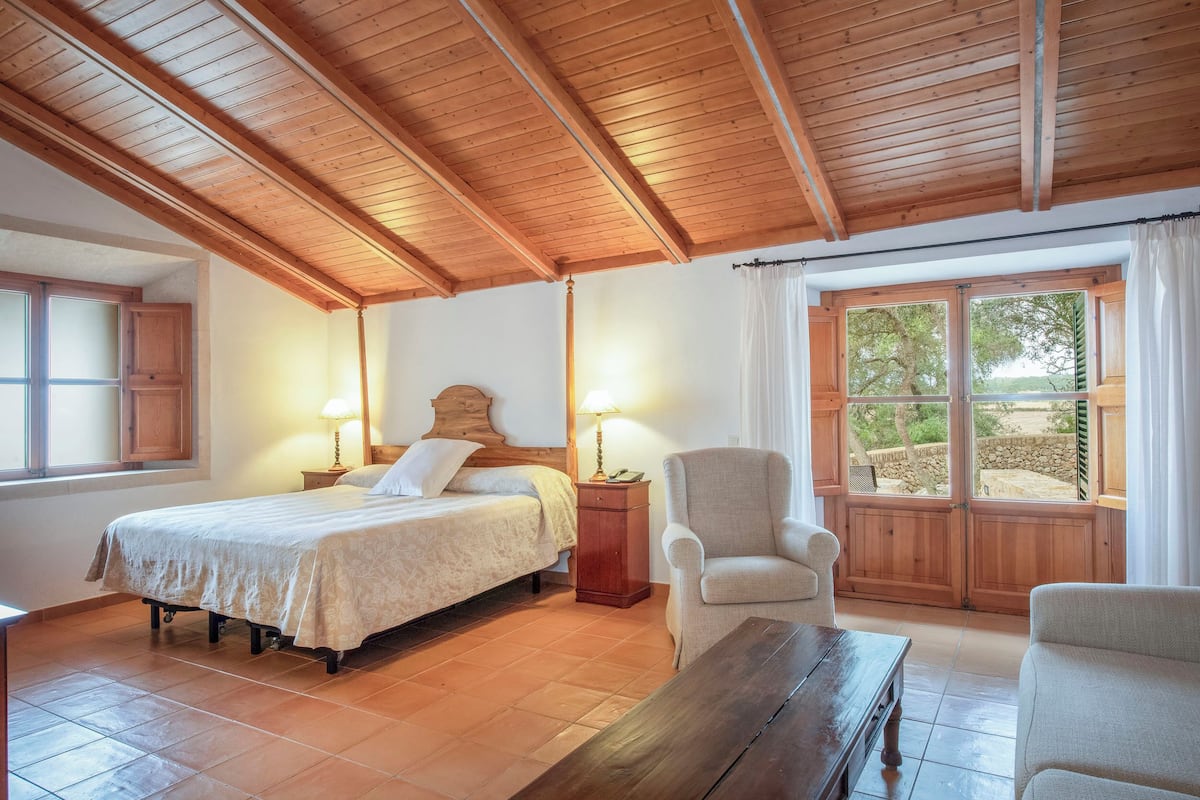 A bedroom with a wooden ceiling, a bed with a headboard, a chair, a nightstand, and a window with a view of trees.