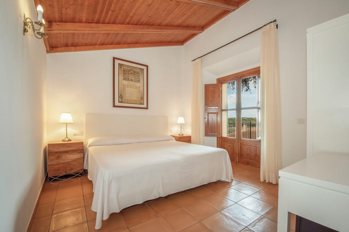 A bedroom with a bed, two bedside lamps, a wooden headboard, a framed picture on the wall, a window with curtains, and a view of the outdoors.