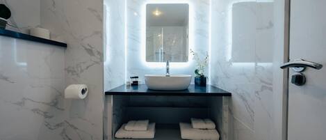 Un baño moderno con lavamanos de mármol, un espejo con iluminación integrada y un estante con toallas.