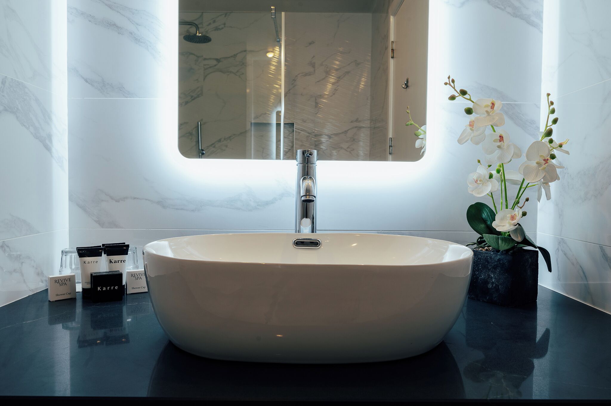 Un bagno con un lavandino bianco grande, uno specchio sopra e un vaso con fiori bianchi sul ripiano.