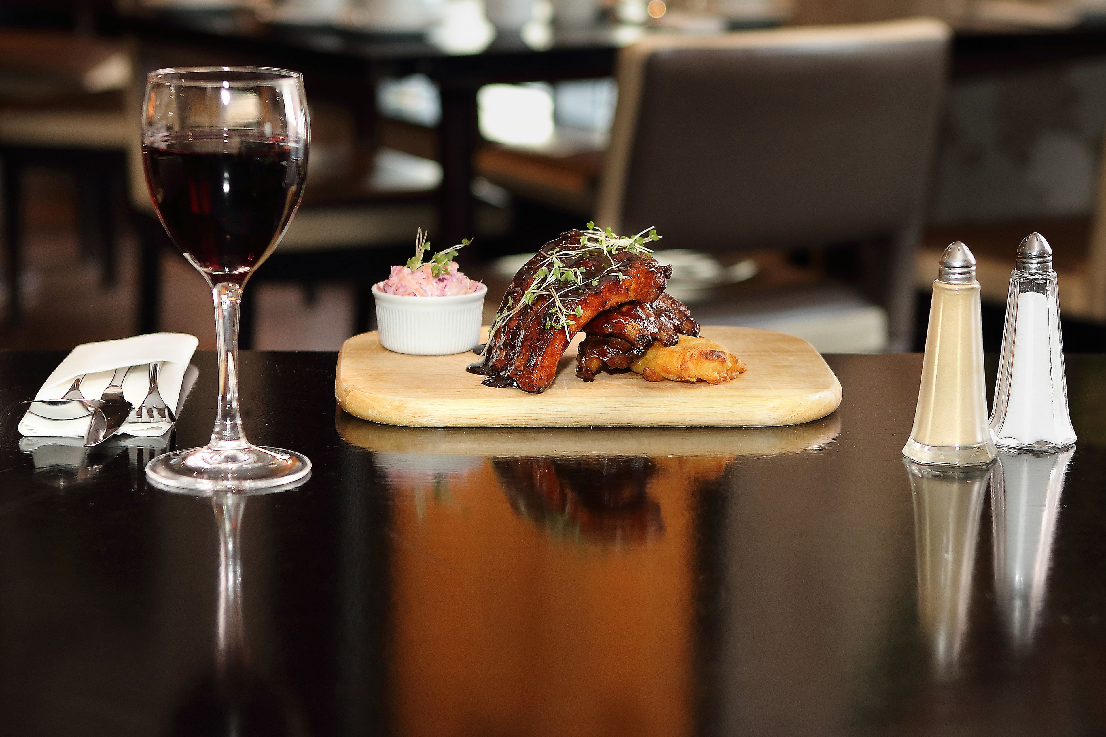 A dining table with a glass of red wine, a plate of ribs garnished with greens, a small bowl of pinkish food, and salt and pepper shakers.