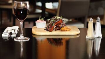 A dining table with a glass of red wine, a plate of ribs garnished with greens, a small bowl of pinkish food, and salt and pepper shakers.