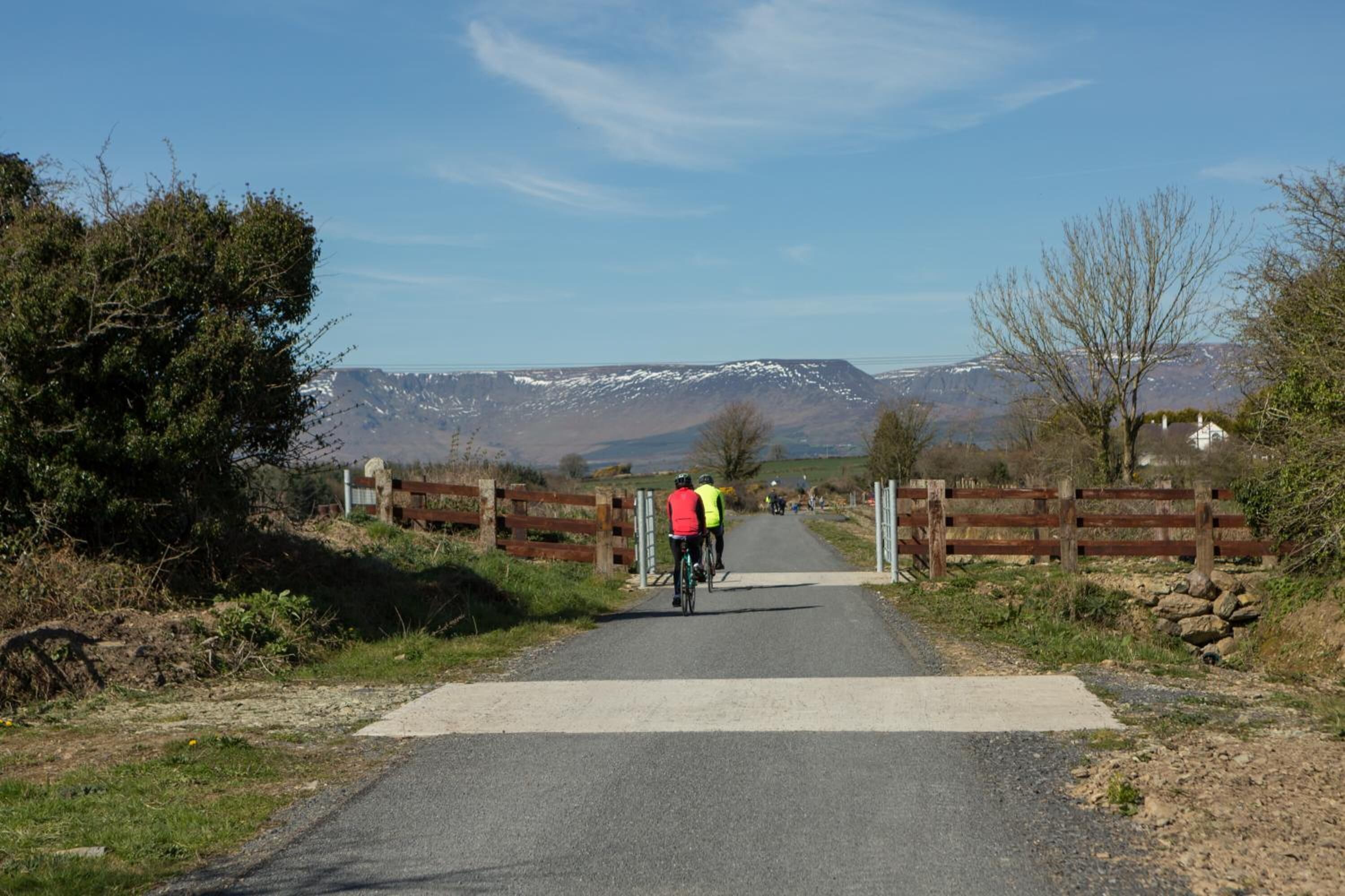 Uma estrada asfaltada com ciclistas, uma cerca de madeira e uma montanha ao fundo.