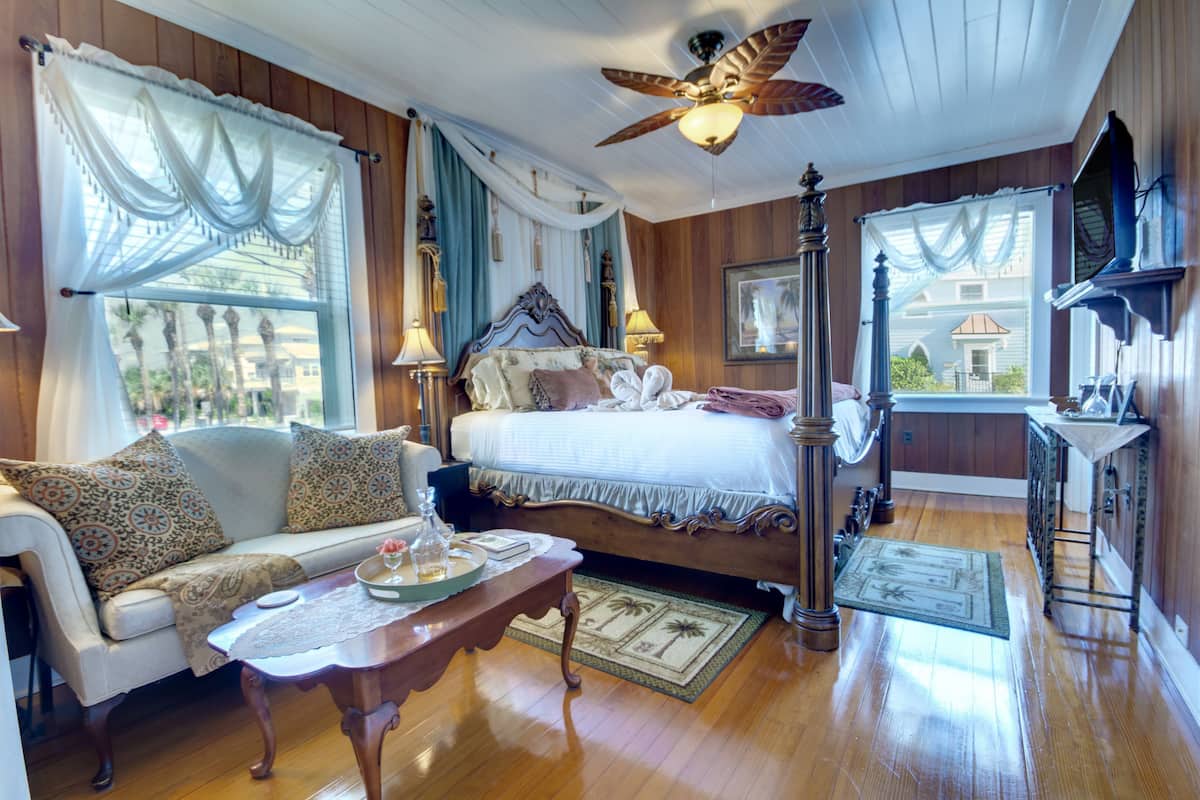 A cozy room with a bed, a sofa, a wooden floor, a ceiling fan, and a window with curtains.
