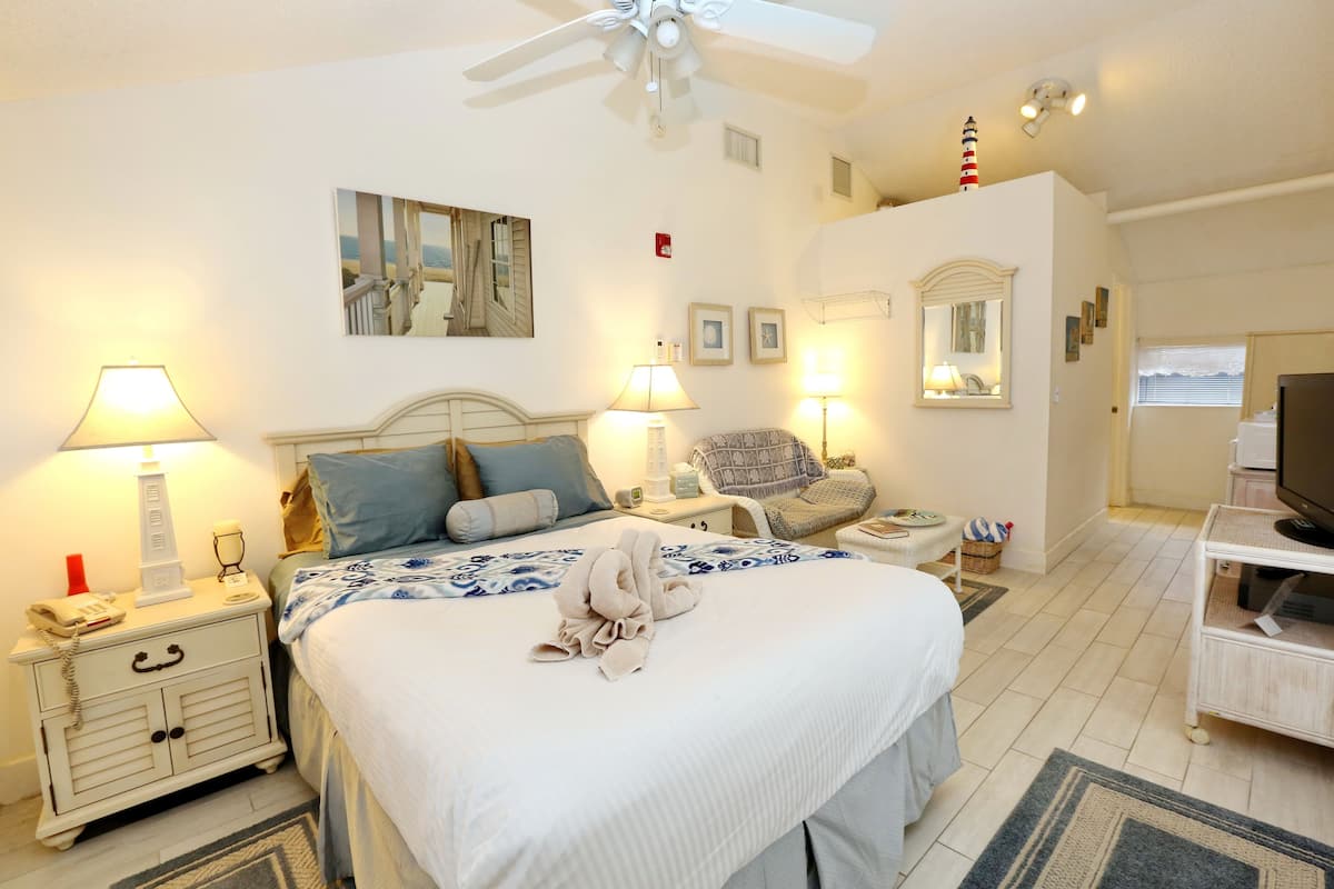 A neatly arranged bedroom with a bed, bedside lamps, a chair, a TV stand, and a ceiling fan.