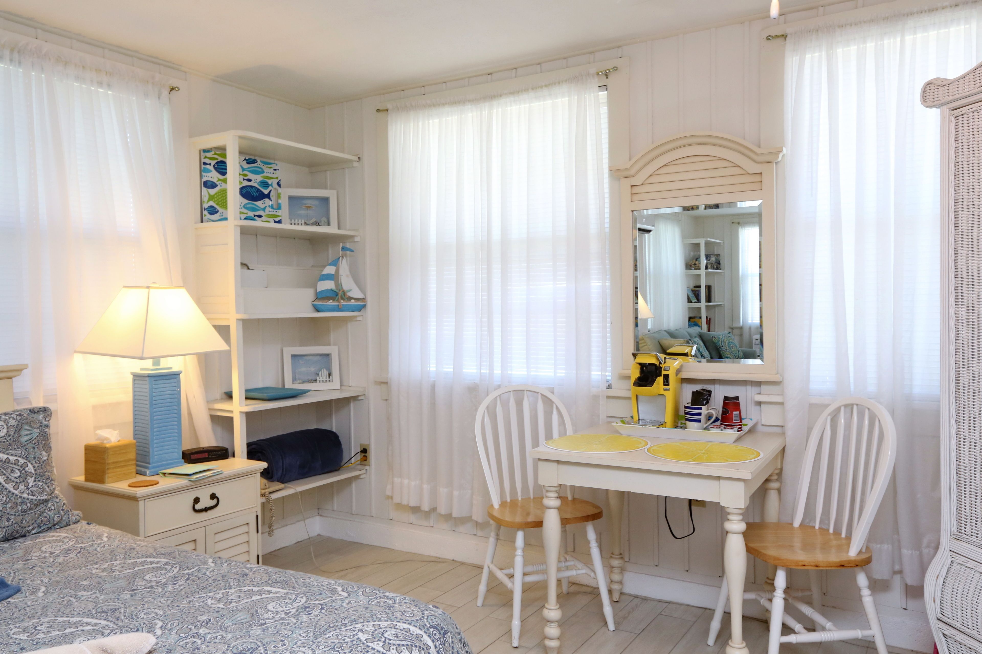 Ein Schlafzimmer mit Bett, Nachttisch, Lampe und Schreibtisch mit Stühlen. Ein Kleiderschrank und ein Spiegel befinden sich an der Wand.