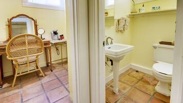 A bathroom with a toilet, sink, and mirror. There is a wicker chair and a small table with a coffee maker. The walls are yellow, and the floor has a tiled pattern.
