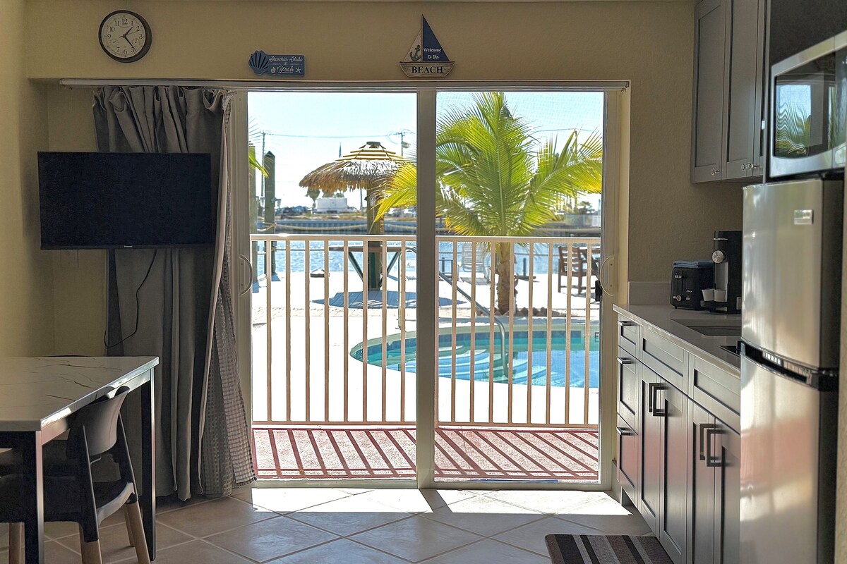 A room with a kitchen, a dining area, a TV, and a sliding glass door leading to a pool area.