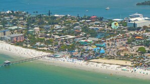 An aerial view of a coastal city with a long pier extending into the sea, numerous buildings, and a beach area with umbrellas and people. - Manatee Bay Inn - Near Fishing Pier Fort Myers Beach (Fort Myers Beach)