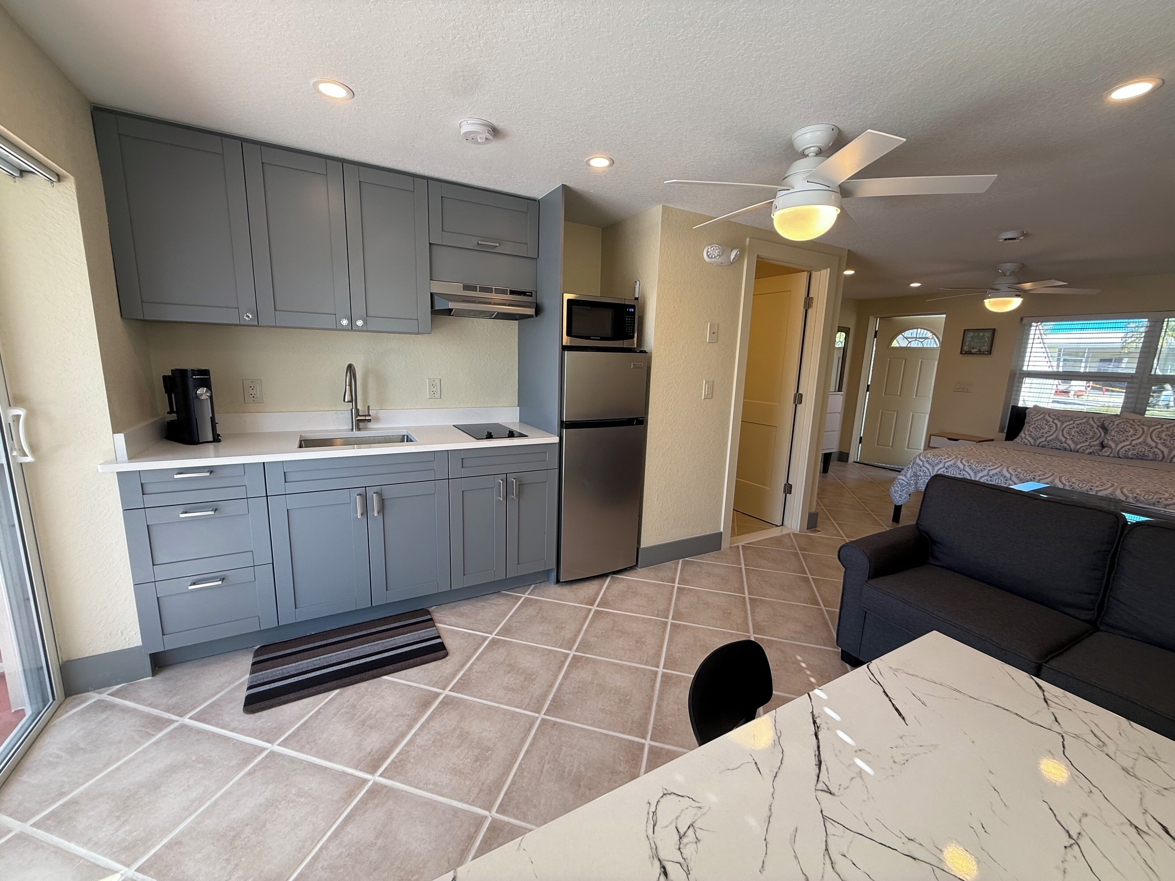A compact kitchen with grey cabinets, a microwave, and a refrigerator. There is a dining area with a marble table and a black chair. A ceiling fan and recessed lighting are visible.