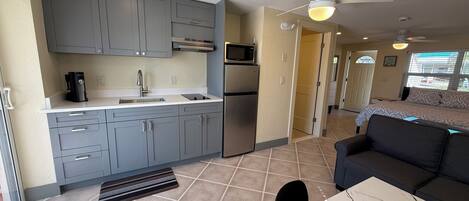 A compact kitchen with grey cabinets, a microwave, and a refrigerator. There is a dining area with a marble table and a black chair. A ceiling fan and recessed lighting are visible.