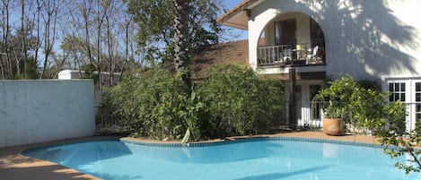 A pool area with lounge chairs, a white building with a balcony, and surrounding greenery.