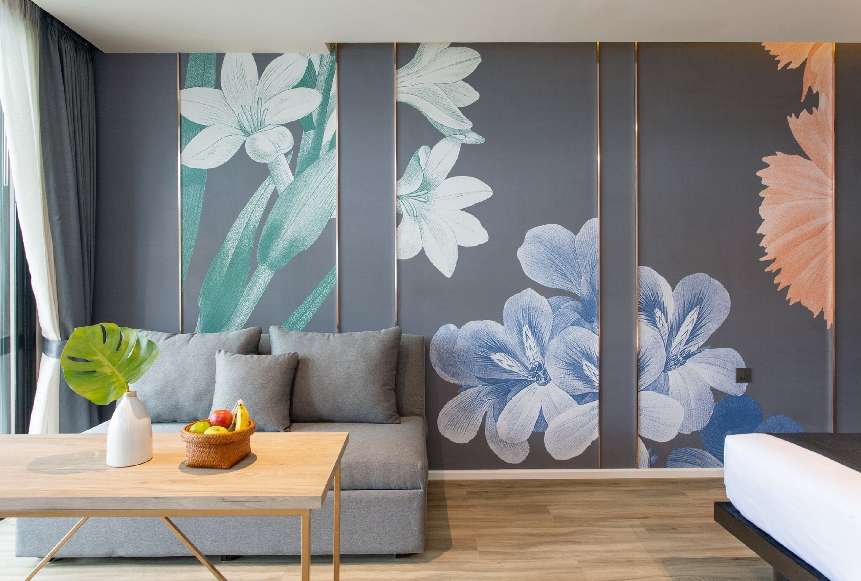 A modern living room with a grey sofa, a wooden coffee table with a fruit basket, and a large wall mural with floral designs.