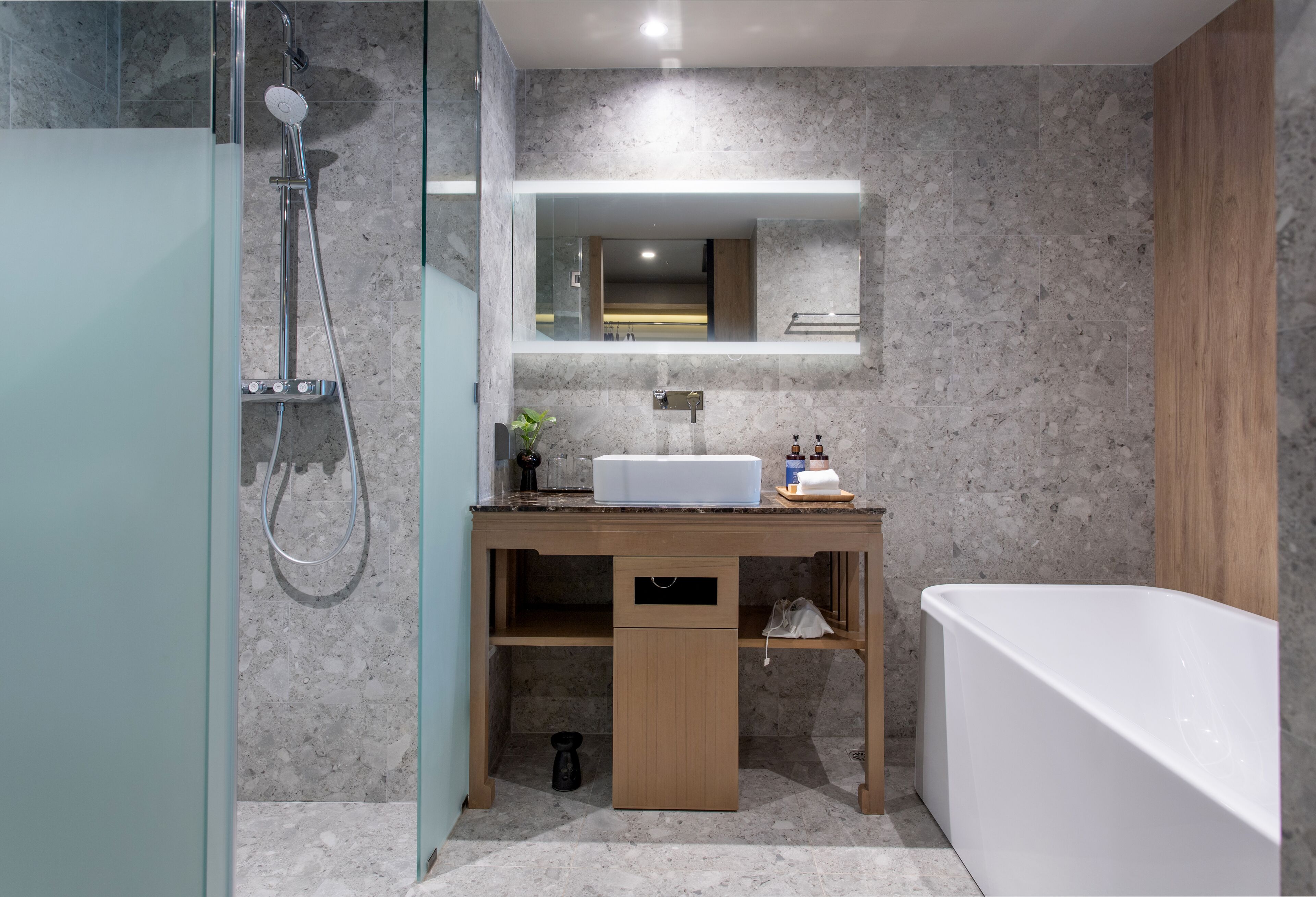 Une salle de bain moderne avec une douche à l’italienne en verre, un meuble-lavabo en bois, une baignoire blanche et un mur recouvert de carreaux de marbre.